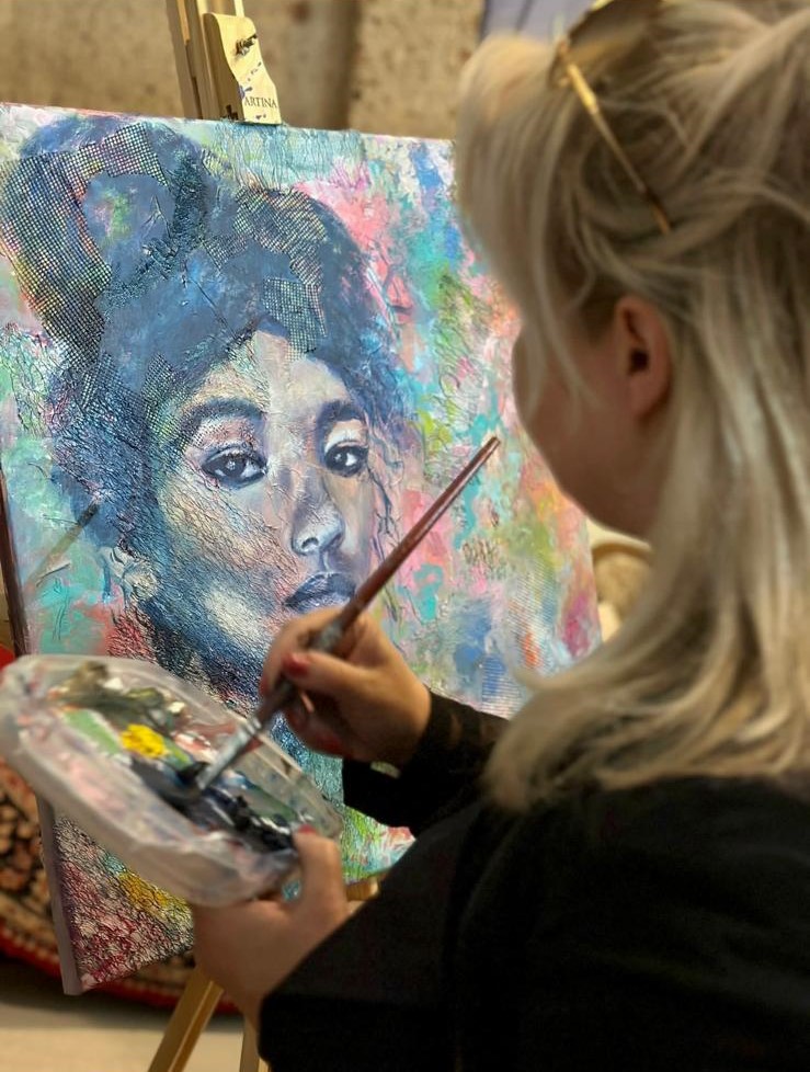 Foto gemaakt over de schouder van een vrouw die bezig is met een pastelkleurig schilderij van een vrouw met een knot. 